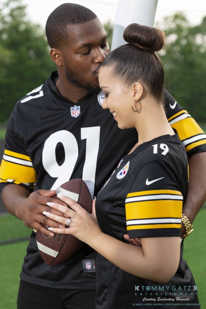 051919-Viona-Kevin-FINALS-Low-RES-WM-47 - TOMMY GATZ ENTERTAINMENT Couple wearing black football jerseys, embracing in an outdoor setting. The man holds a football as he leans in toward the woman.