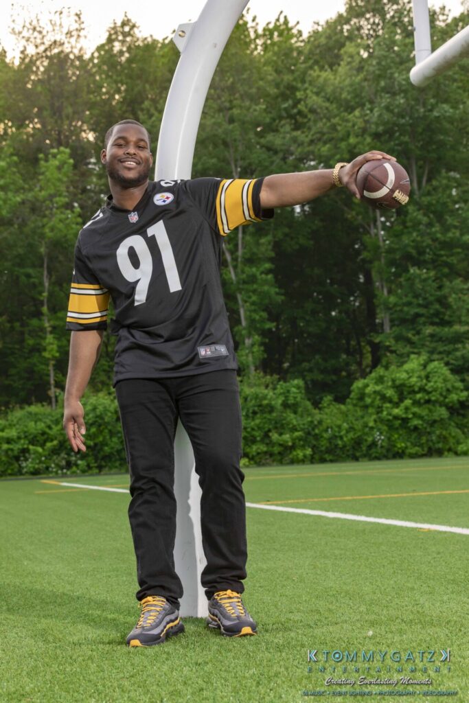 051919-Viona-Kevin-FINALS-Low-RES-WM-48 - TOMMY GATZ ENTERTAINMENT A person wearing a black and yellow football jersey poses with a football under an upright post on a green field.