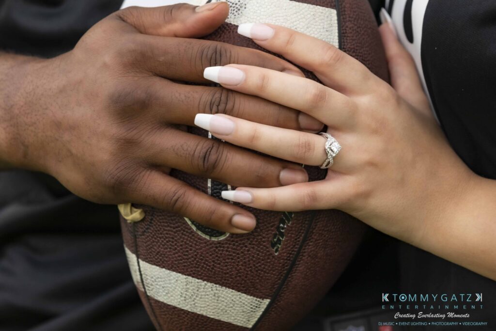 051919-Viona-Kevin-FINALS-Low-RES-WM-49 - TOMMY GATZ ENTERTAINMENT Two hands with manicured nails and a diamond ring rest on a football, showcasing diversity and engagement.