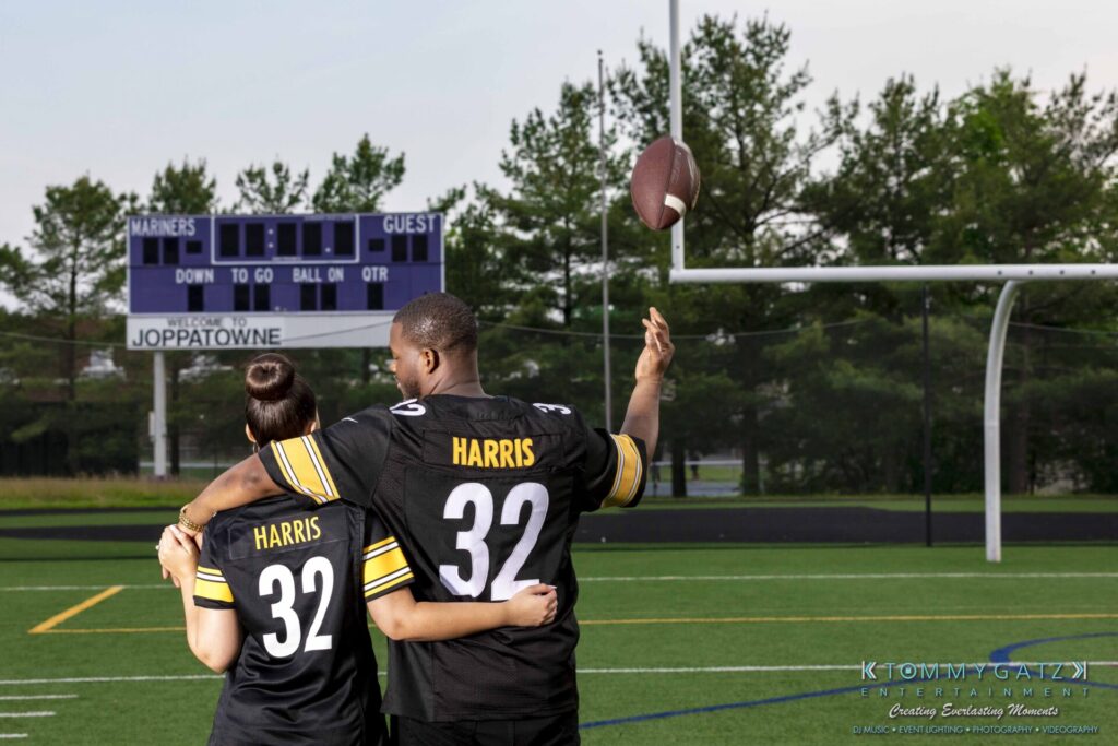 051919-Viona-Kevin-FINALS-Low-RES-WM-51 - TOMMY GATZ ENTERTAINMENT Two people in matching jerseys, labeled "Harris 32," stand on a football field. One person is holding a football with an outstretched arm. A scoreboard and goalpost are visible in the background.