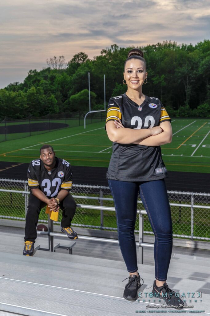 051919-Viona-Kevin-FINALS-Low-RES-WM-52 - TOMMY GATZ ENTERTAINMENT Two people in matching sports jerseys are at a stadium. One stands confidently on bleachers, while the other sits behind, looking toward the camera. A football field is in the background.
