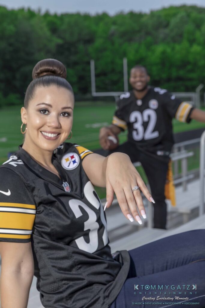 051919-Viona-Kevin-FINALS-Low-RES-WM-53 - TOMMY GATZ ENTERTAINMENT A woman shows her engagement ring while sitting in a stadium, with a man holding a football in the background. Both are wearing football jerseys with the number 32.