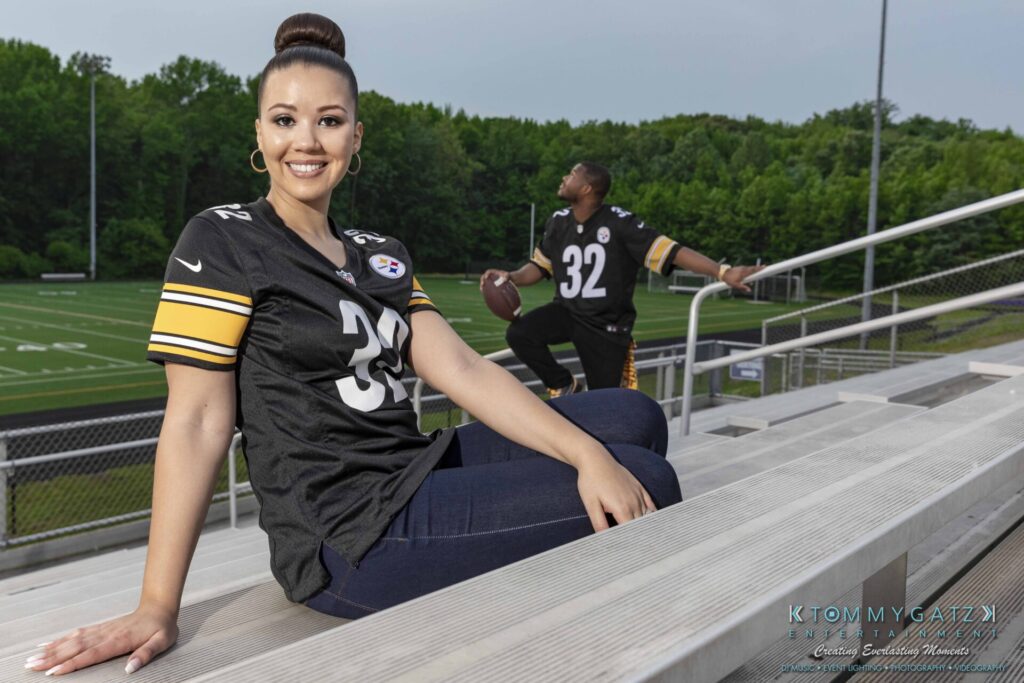 051919-Viona-Kevin-FINALS-Low-RES-WM-54 - TOMMY GATZ ENTERTAINMENT Two people wearing matching sports jerseys sit on bleachers near a football field. One person is seated and smiling at the camera, while the other holds a football and looks toward the field.