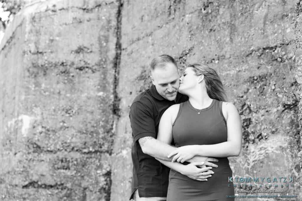 063019-Melanie-Rob-FINALS-Low-Res-WM-11 - TOMMY GATZ ENTERTAINMENT A couple embracing; the woman leans in to kiss the man's cheek. They're standing against a textured stone wall.
