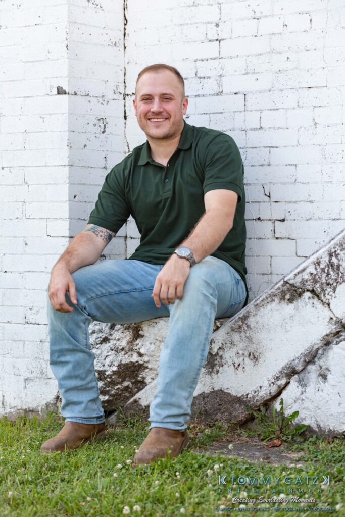 063019-Melanie-Rob-FINALS-Low-Res-WM-12 - TOMMY GATZ ENTERTAINMENT Smiling person in a green polo shirt and jeans sits on a concrete ledge against a white brick wall outdoors.