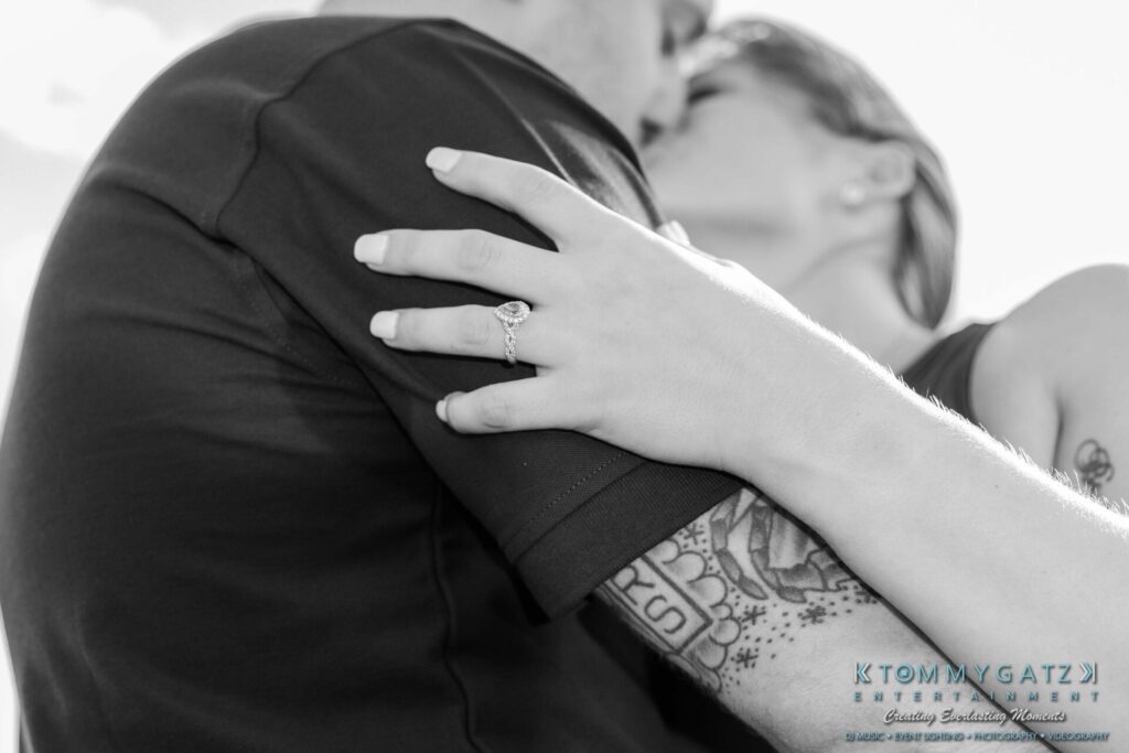 063019-Melanie-Rob-FINALS-Low-Res-WM-17 - TOMMY GATZ ENTERTAINMENT Close-up of a couple kissing, showcasing a diamond engagement ring on the woman's hand resting on the man's shoulder.