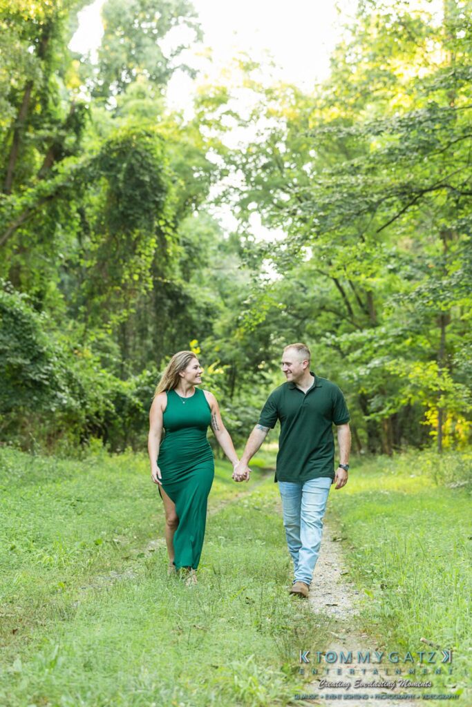063019-Melanie-Rob-FINALS-Low-Res-WM-19 - TOMMY GATZ ENTERTAINMENT A man and woman hold hands walking on a grassy path in a wooded area. Both are dressed in green outfits, with the woman in a long dress and the man in a polo shirt and jeans.