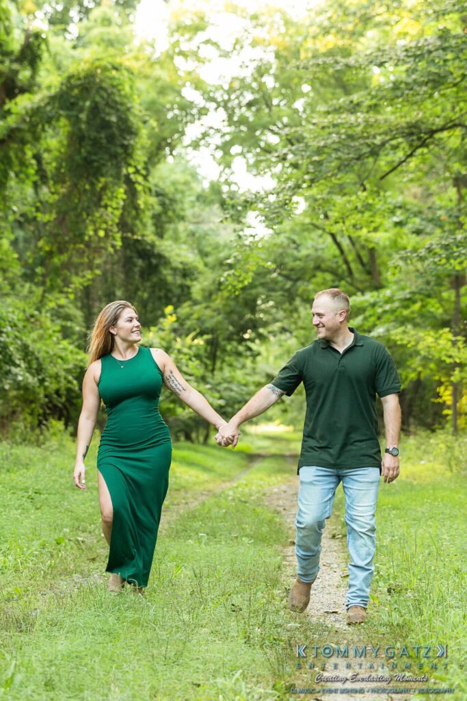 063019-Melanie-Rob-FINALS-Low-Res-WM-20 - TOMMY GATZ ENTERTAINMENT A couple holding hands and walking on a grassy path in a lush green forest, both wearing green outfits.