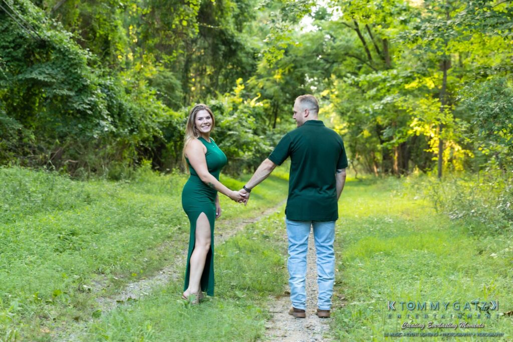 063019-Melanie-Rob-FINALS-Low-Res-WM-22 - TOMMY GATZ ENTERTAINMENT Couple holding hands and walking on a grassy path in a lush, green forest. The woman wears a green dress, and the man is in a green shirt and jeans.