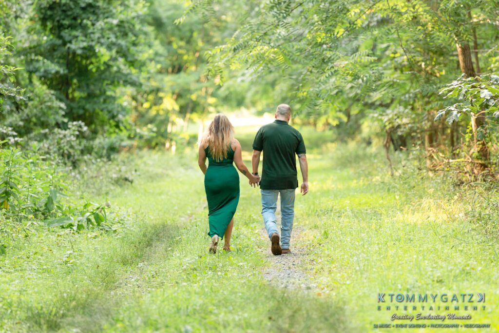 063019-Melanie-Rob-FINALS-Low-Res-WM-23 - TOMMY GATZ ENTERTAINMENT A couple holding hands walks along a grassy trail surrounded by trees.