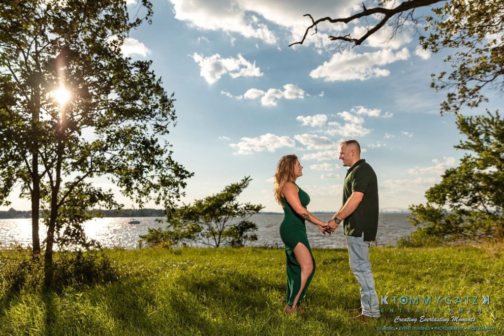 063019-Melanie-Rob-FINALS-Low-Res-WM-25 - TOMMY GATZ ENTERTAINMENT A couple holds hands by a sunlit waterfront, surrounded by grass and trees, with a clear sky above.