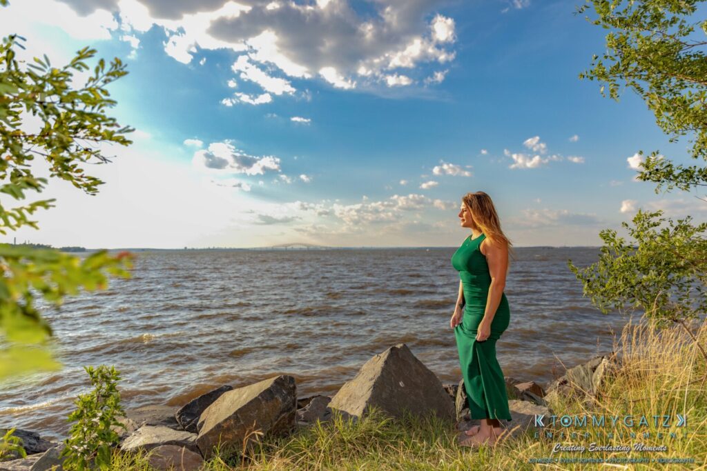 063019-Melanie-Rob-FINALS-Low-Res-WM-27 - TOMMY GATZ ENTERTAINMENT Woman in a green dress stands on rocks by a lake, looking at the horizon under a partly cloudy sky. Trees and grass are in the foreground.