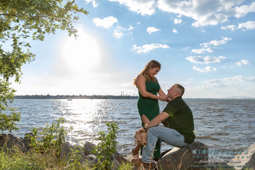 063019-Melanie-Rob-FINALS-Low-Res-WM-29 - TOMMY GATZ ENTERTAINMENT A couple poses by the rocky shoreline under a bright, sunny sky; the woman stands while the man kneels, holding her hand.