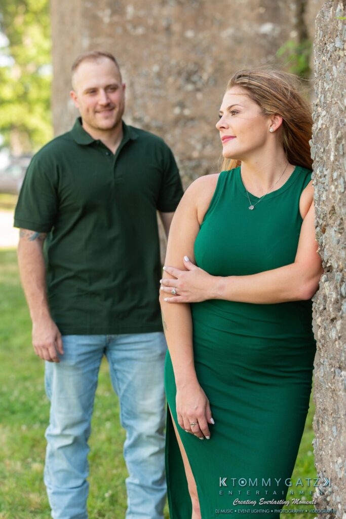 063019-Melanie-Rob-FINALS-Low-Res-WM-3 - TOMMY GATZ ENTERTAINMENT A woman in a green dress leans against a stone wall, looking to the side. A man in a dark green shirt and jeans stands behind her, smiling. They are outdoors.