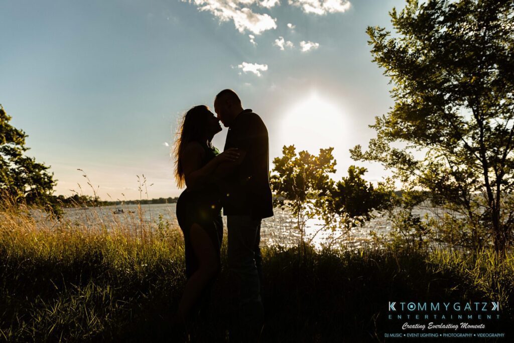 063019-Melanie-Rob-FINALS-Low-Res-WM-33 - TOMMY GATZ ENTERTAINMENT Silhouetted couple stands in the grass by a lake at sunset, with trees and water visible in the background.