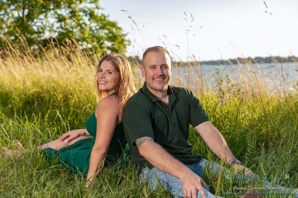 063019-Melanie-Rob-FINALS-Low-Res-WM-36 - TOMMY GATZ ENTERTAINMENT A man and a woman sit on the grass near a body of water, smiling and facing the camera.