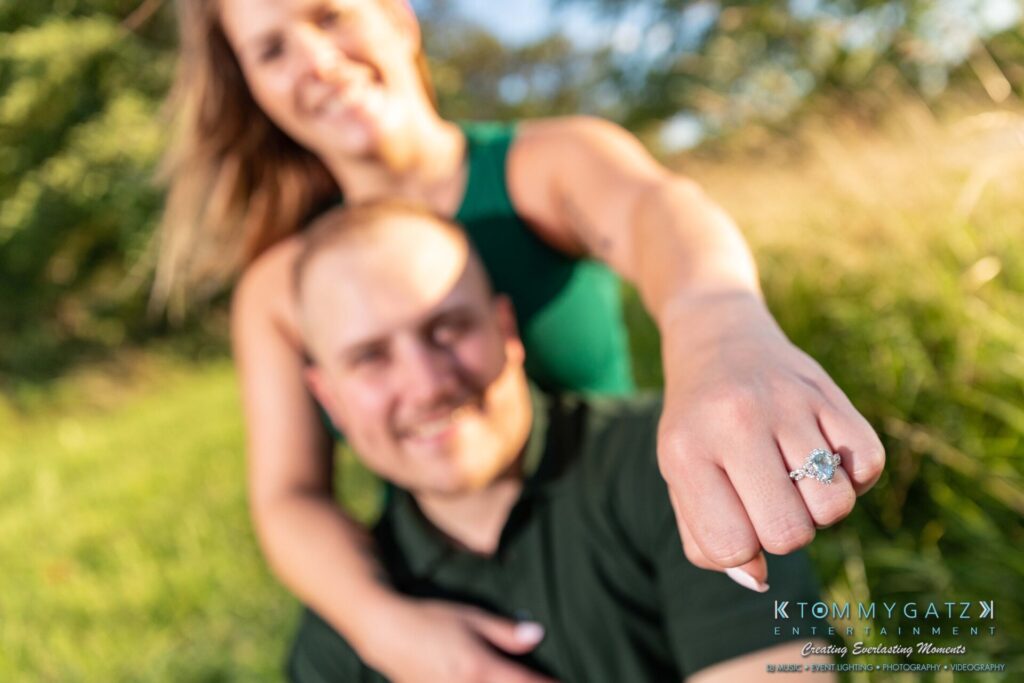 063019-Melanie-Rob-FINALS-Low-Res-WM-40 - TOMMY GATZ ENTERTAINMENT A couple sits outdoors, with the woman in focus showing her engagement ring. She is smiling, while the man appears slightly blurred in the background.