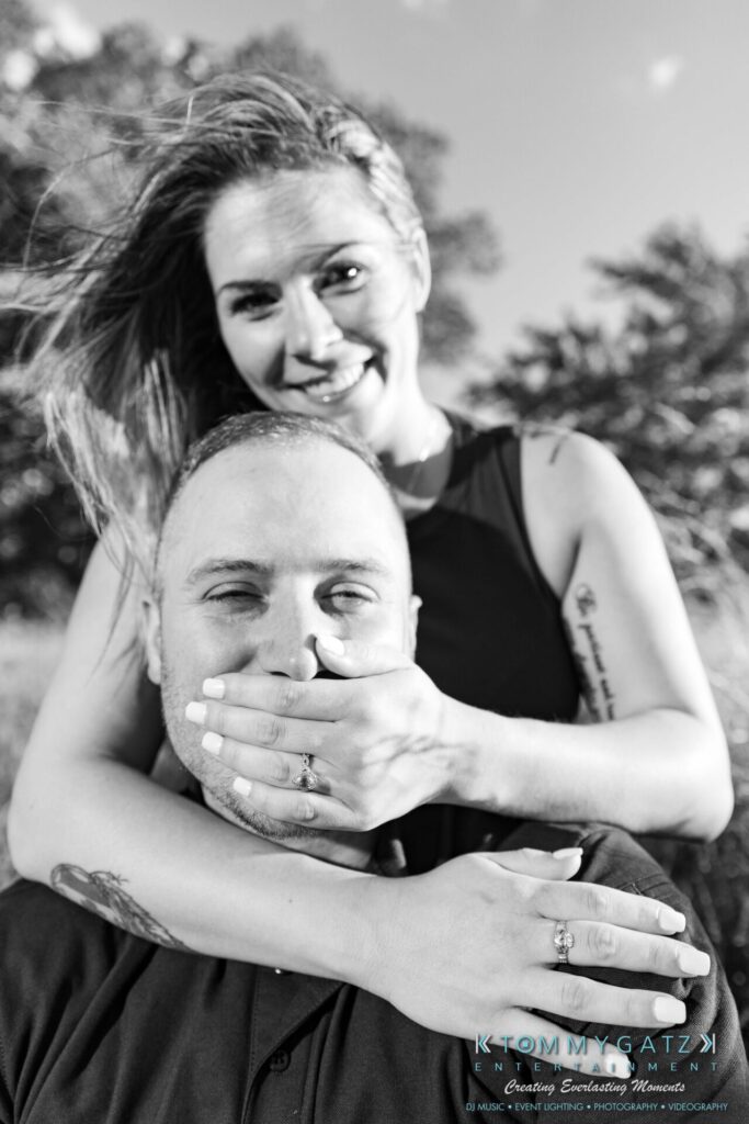 063019-Melanie-Rob-FINALS-Low-Res-WM-41 - TOMMY GATZ ENTERTAINMENT A black and white photo of a woman playfully covering a man's mouth with her hand. Both are outdoors with a tree and sky in the background.