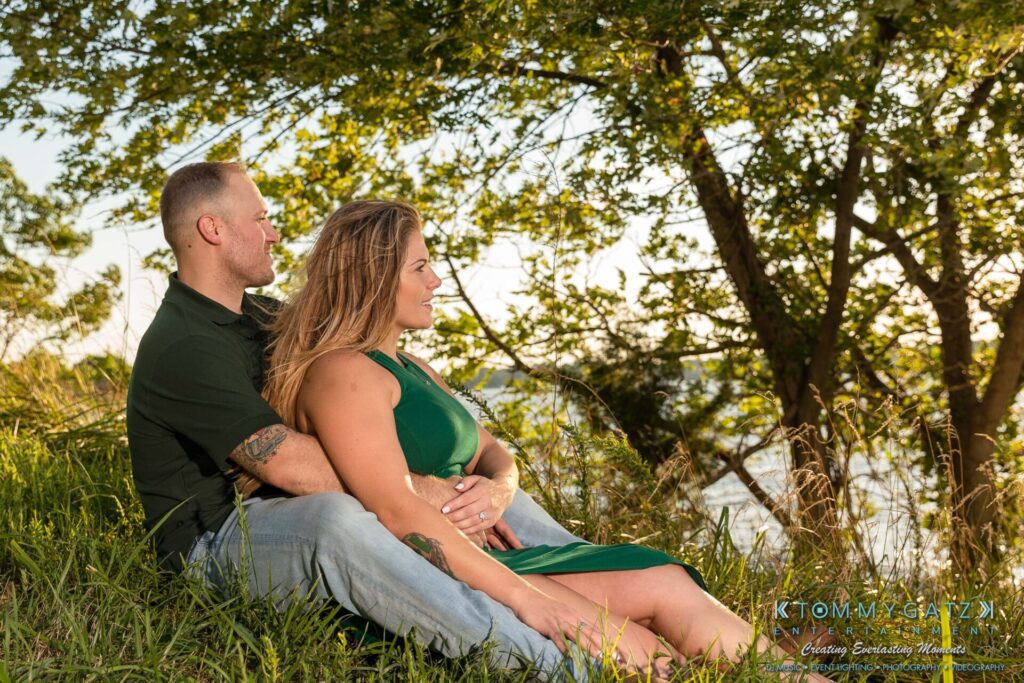 063019-Melanie-Rob-FINALS-Low-Res-WM-42 - TOMMY GATZ ENTERTAINMENT A couple sits closely on a grassy hill, gazing into the distance, with trees and sunlight in the background.