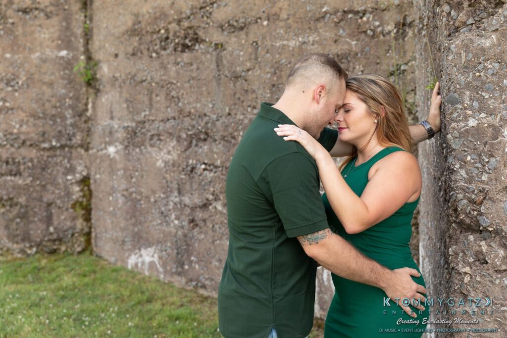 063019-Melanie-Rob-FINALS-Low-Res-WM-6 - TOMMY GATZ ENTERTAINMENT A couple embraces affectionately against a stone wall. Both are wearing green attire, and the setting is outdoors with grass visible nearby.