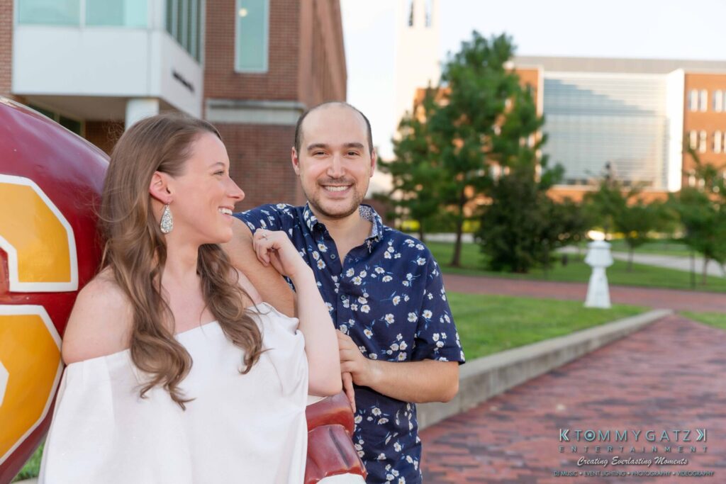 A woman and a man smiling in a park setting, leaning against a large red and yellow sculpture. Brick paths and buildings are visible in the background.