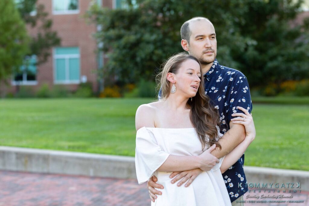 A couple embracing outdoors; the woman is wearing an off-shoulder white dress, and the man is in a patterned shirt. They stand in front of a lawn and trees.