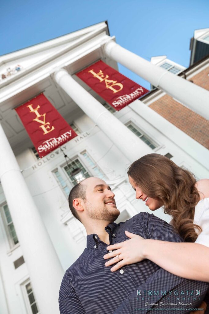 A couple stands in front of a building with red banners labeled "LIVE" and "SANCTIFY," smiling at each other.