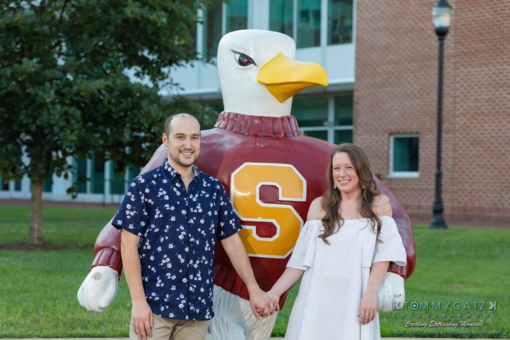 A couple holds hands in front of a large eagle statue wearing a maroon sweater with a yellow "S" on it. They are outdoors near a brick building and a tree.