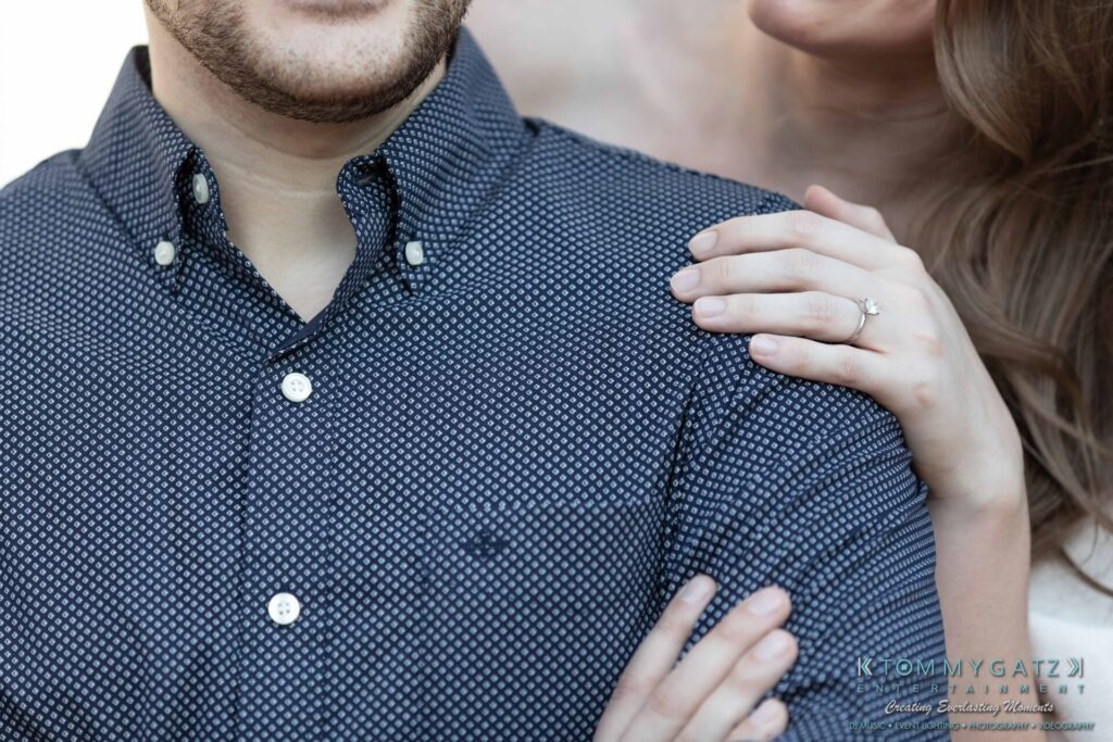 Close-up of a couple, with one person's arm around the other's shoulder. The person in the back is wearing an engagement ring.