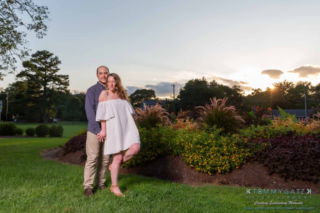 A couple stands together in a garden at sunset. The woman is wearing a white dress, and the man is in a blue shirt and khaki pants.