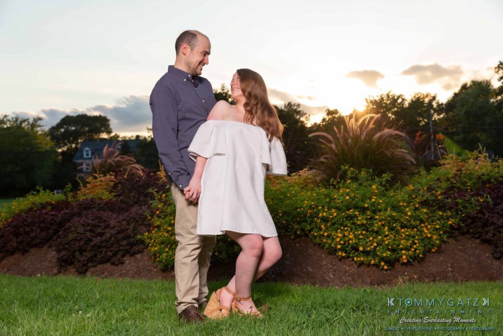 A couple stands outside holding hands and smiling at each other. The sun sets behind them, casting a warm glow.
