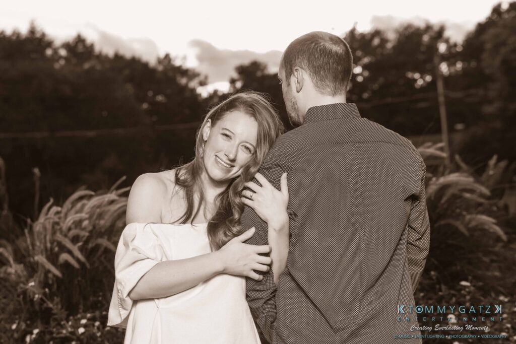 A woman in a white dress leans on a man in a dark shirt, smiling at the camera. They stand outdoors with trees in the background.