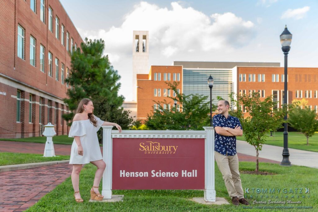 Two people stand on either side of a sign for Henson Science Hall at Salisbury University, with brick buildings and a clock tower in the background.