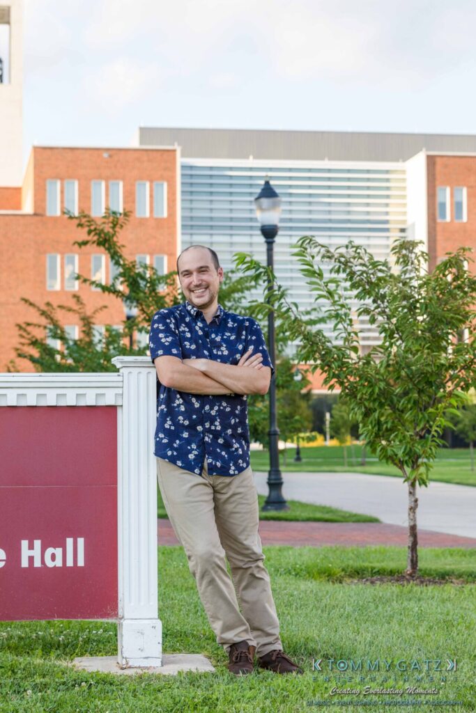 A person in a floral shirt and khaki pants leans against a sign on a green lawn with a brick building in the background.