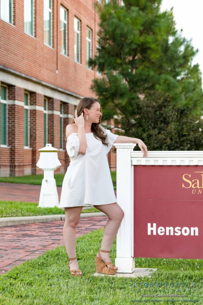 A woman in a white dress poses next to a campus sign outdoors, smiling and placing one hand on her hip.