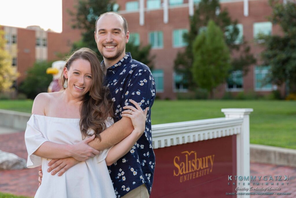 A couple stands smiling in front of a Salisbury University sign with brick buildings and greenery in the background.