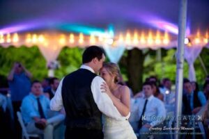 A couple shares a dance at an outdoor wedding reception under a tent, with seated guests watching.