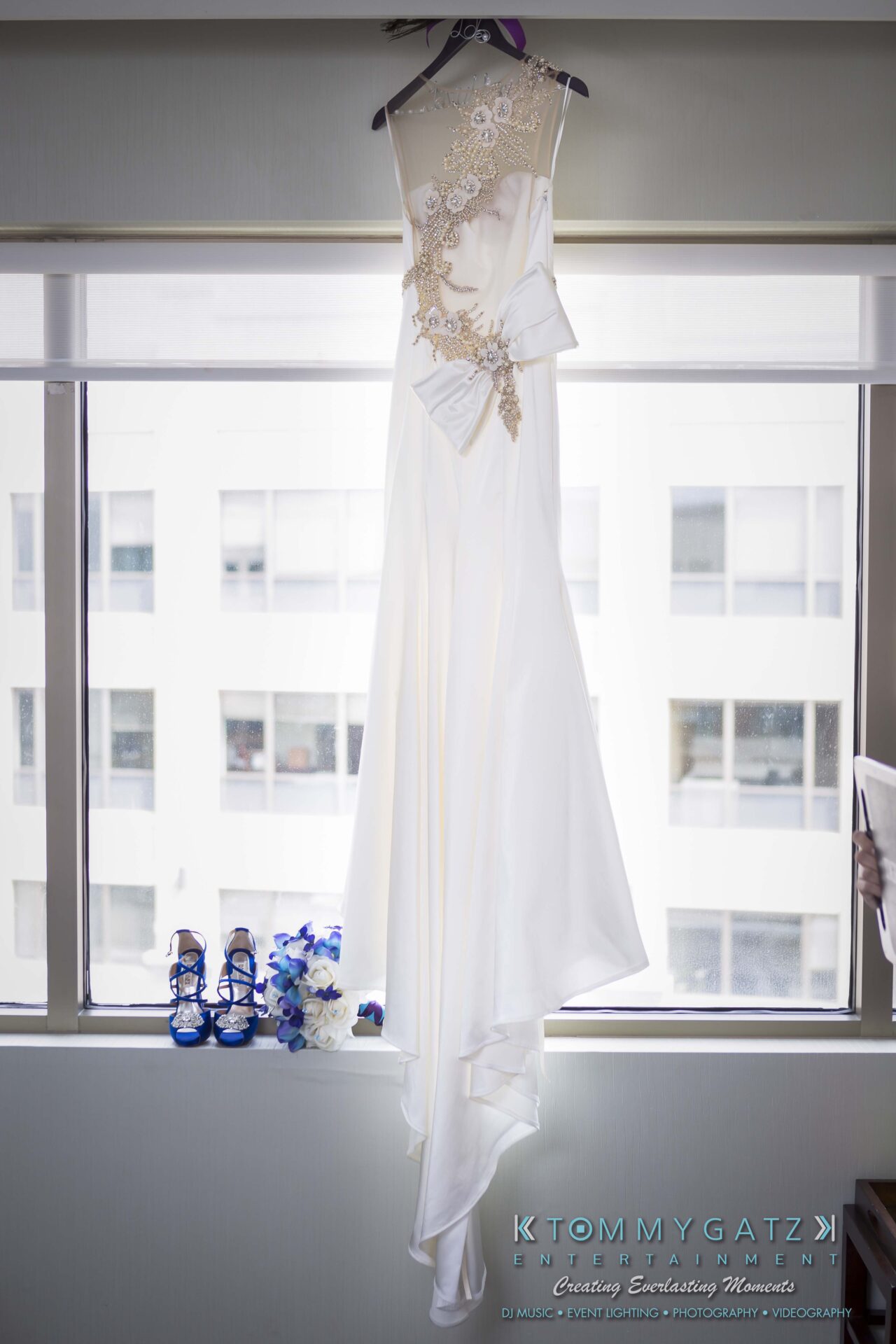 021 - TOMMY GATZ ENTERTAINMENT A white wedding dress with floral lace detail hangs in front of a window. Blue heels and a bouquet are placed on the windowsill below.