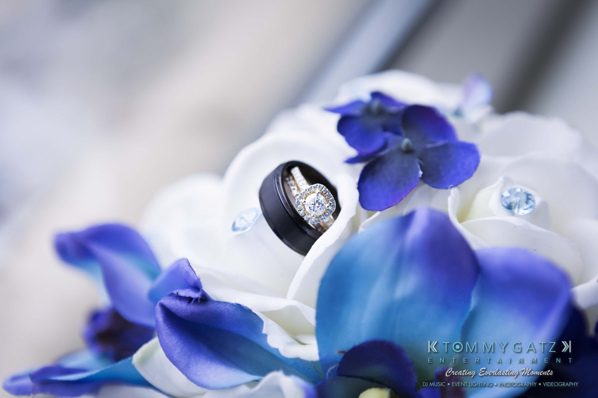031 - TOMMY GATZ ENTERTAINMENT Wedding rings on a white rose bouquet with blue and purple flowers in the background.