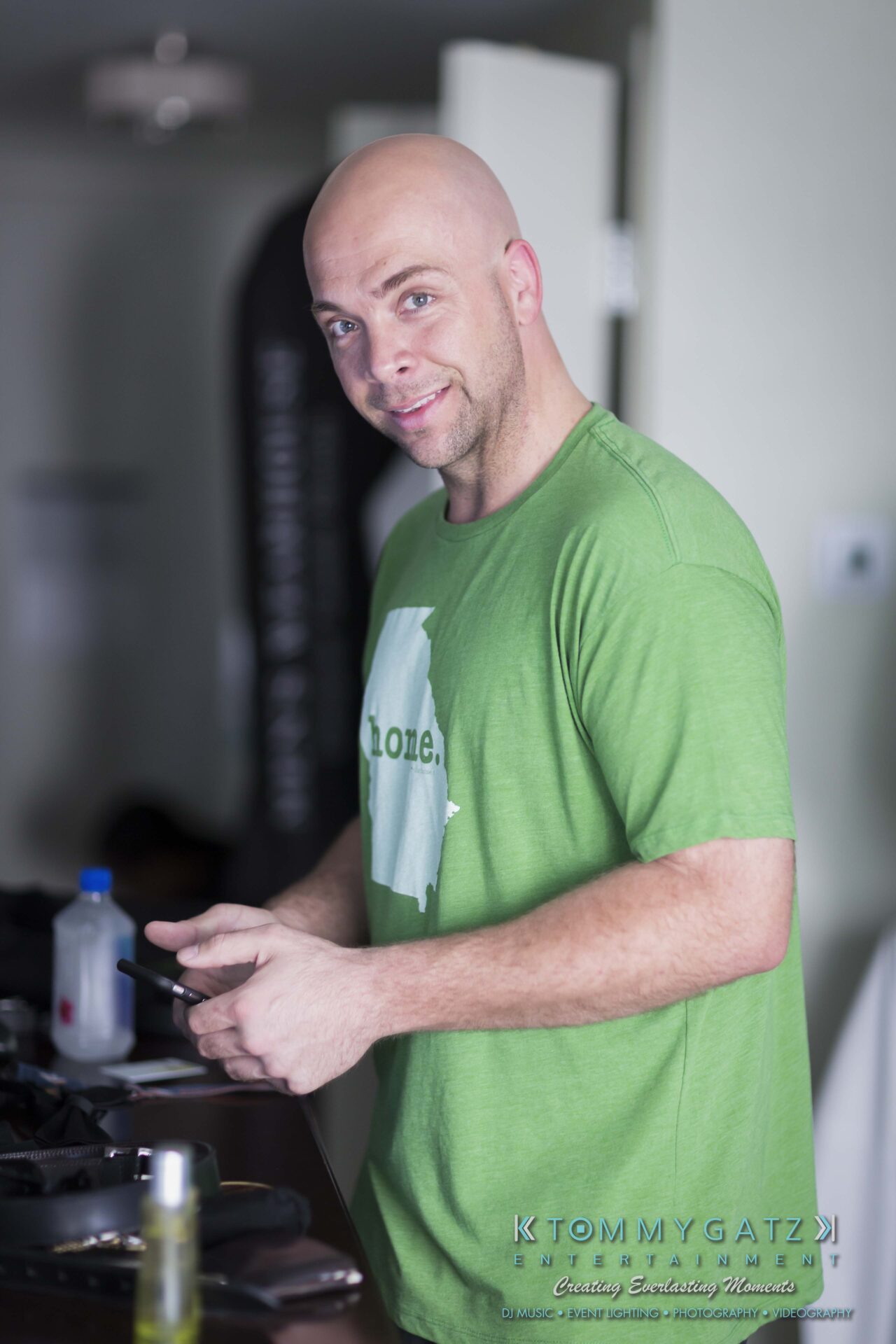 051 - TOMMY GATZ ENTERTAINMENT A man in a green t-shirt stands indoors, holding a phone and smiling at the camera.