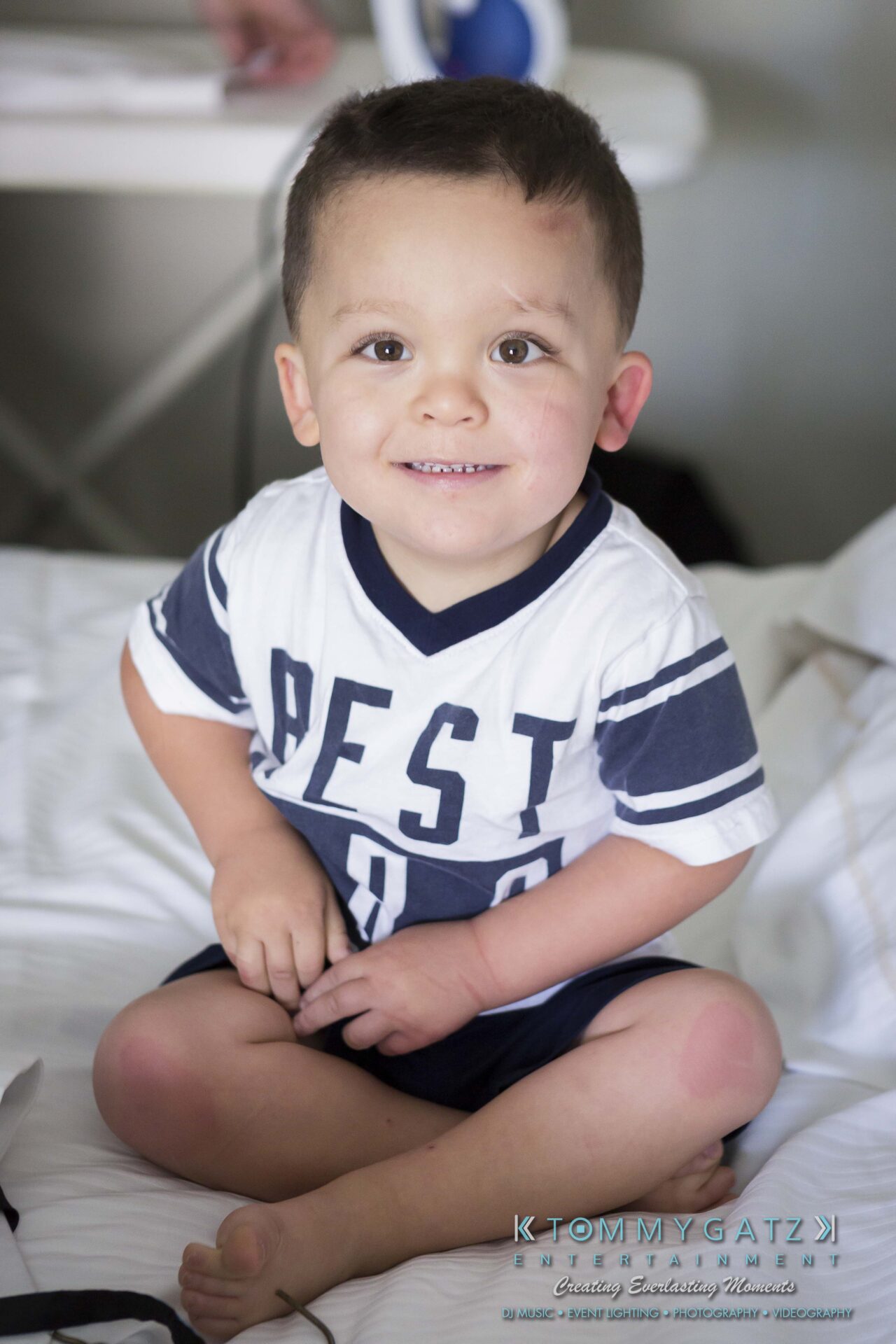 061 - TOMMY GATZ ENTERTAINMENT A young boy sitting on a bed, smiling at the camera, wearing a striped shirt with "BEST" printed on it.