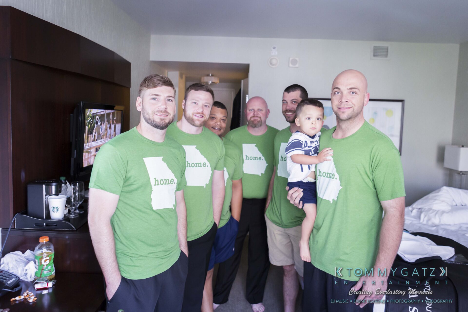 081 - TOMMY GATZ ENTERTAINMENT A group of six men in matching green t-shirts stands together in a room. One man holds a small child. A few drinks are visible in the foreground.