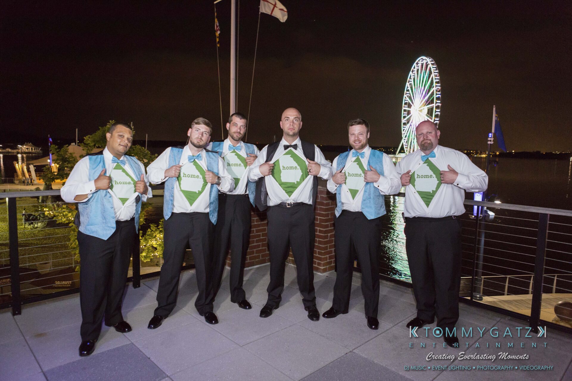103 - TOMMY GATZ ENTERTAINMENT Six men in formal attire stand on a balcony at night, revealing T-shirts with “home” printed on them under their vests. A Ferris wheel and water are in the background.