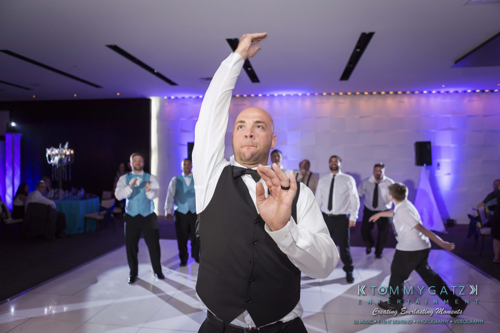 107 - TOMMY GATZ ENTERTAINMENT A bald man in a vest and tie energetically dances on a white floor, with people dressed in formal attire watching and cheering in the background.