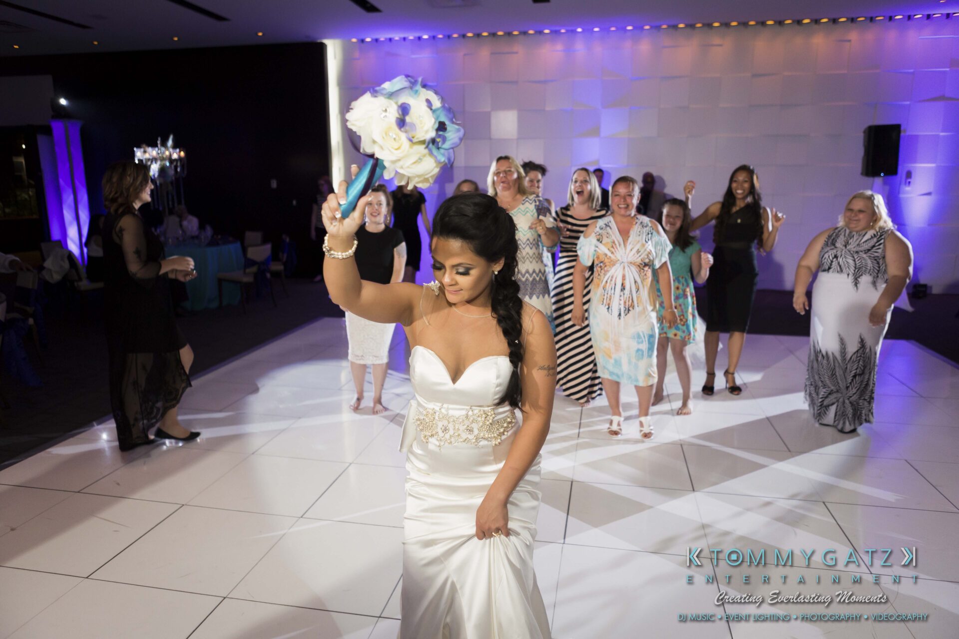 108 - TOMMY GATZ ENTERTAINMENT A bride in a white dress throws a bouquet over her shoulder at a wedding reception, while guests in various outfits stand behind her, eagerly reaching for the bouquet.
