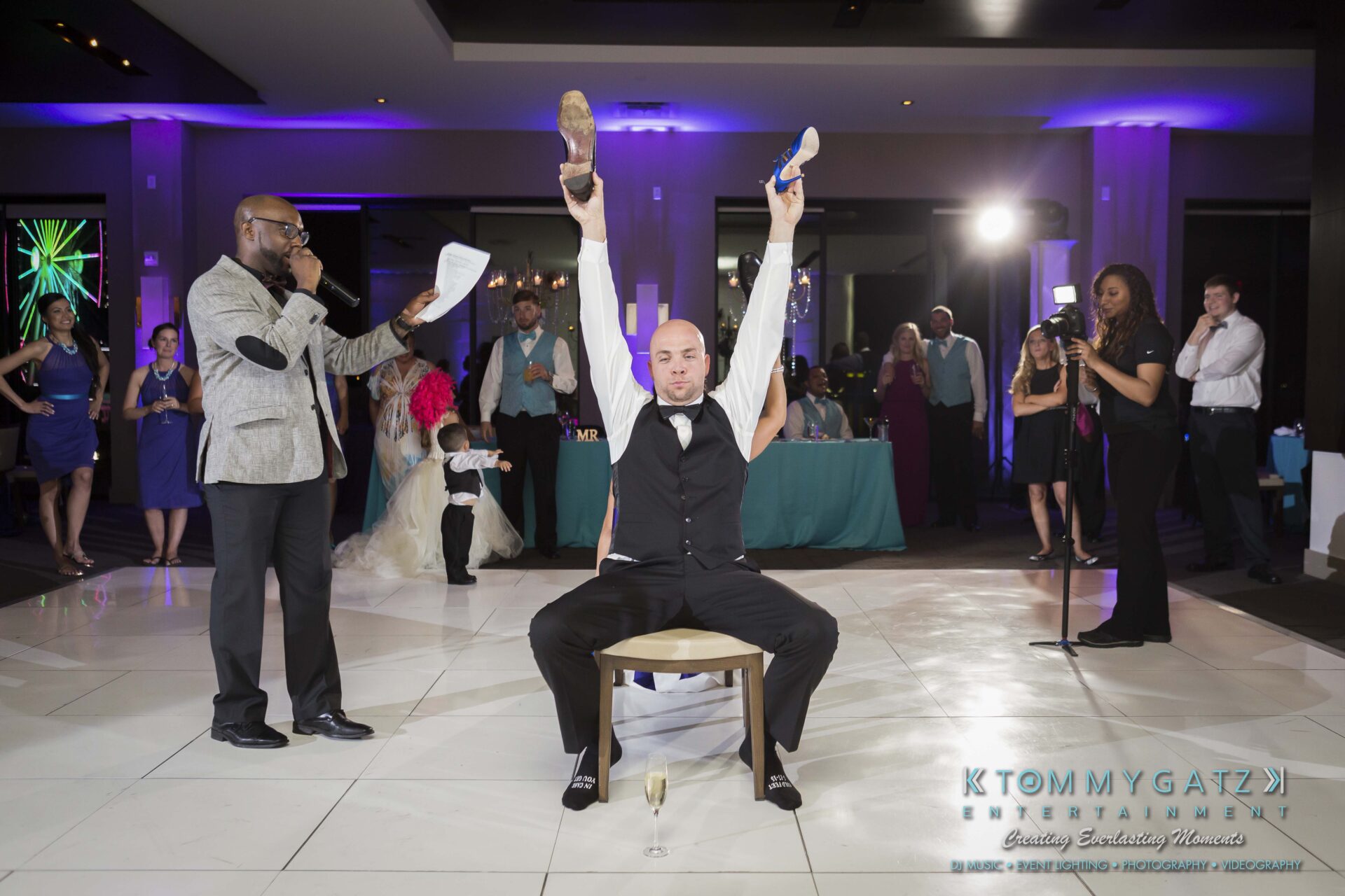 112 - TOMMY GATZ ENTERTAINMENT Man participating in a wedding shoe game, holding a shoe in each hand. An emcee speaks into a microphone, while guests watch.