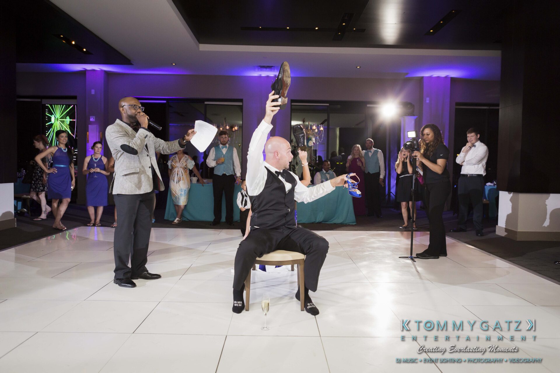 113 - TOMMY GATZ ENTERTAINMENT A man sitting on a chair lifts a shoe, while another man beside him speaks into a microphone at an indoor event. People in the background are observing the scene.
