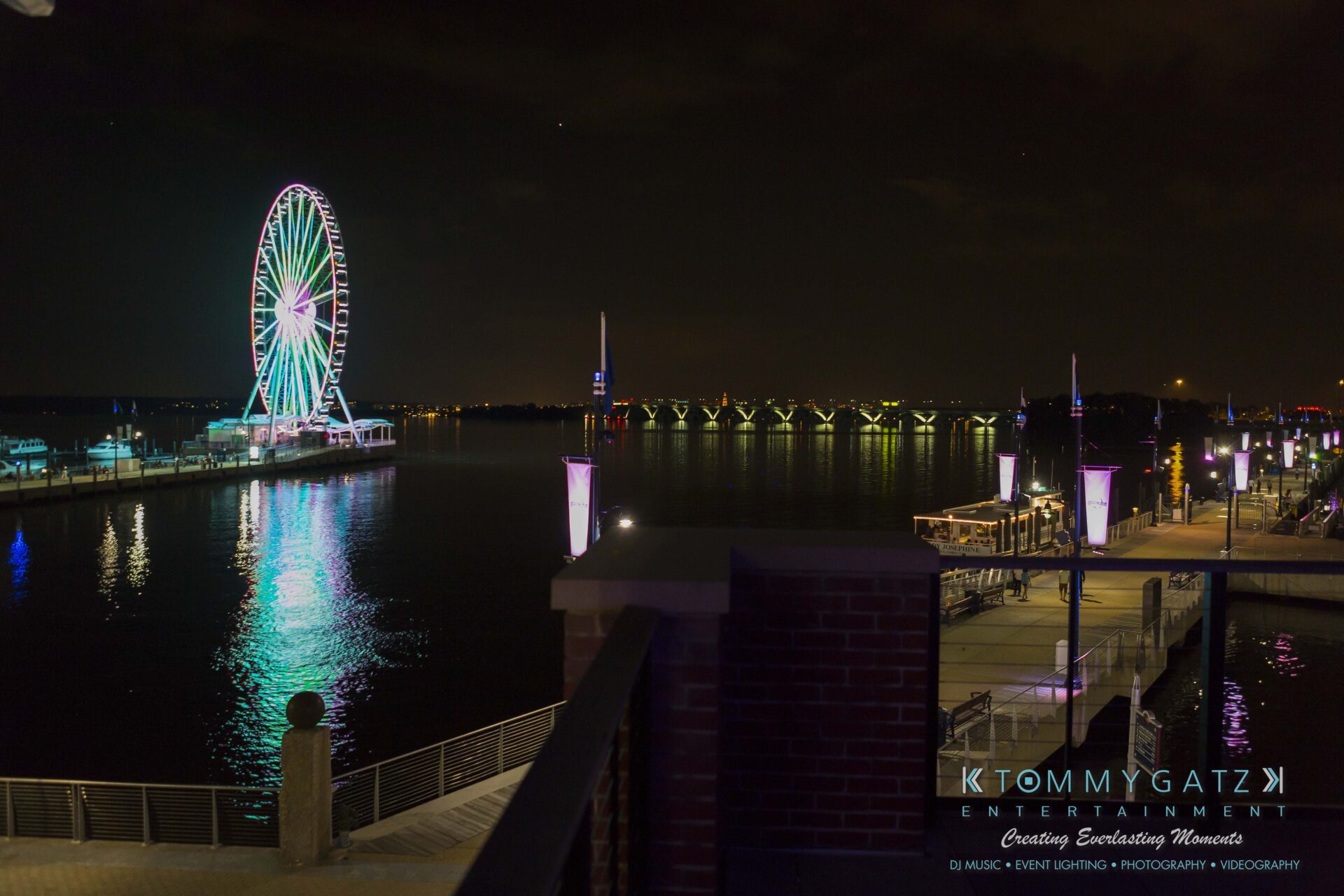 116 - TOMMY GATZ ENTERTAINMENT Night view of a waterfront with a brightly lit Ferris wheel, illuminated walkway, and reflections on the water.