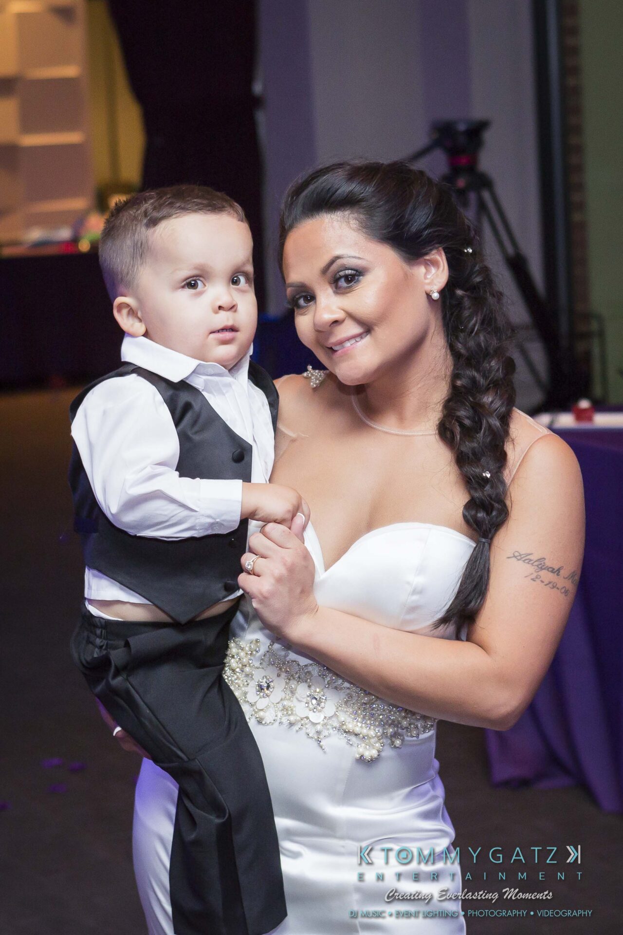 117 - TOMMY GATZ ENTERTAINMENT A woman in a white dress holds a young boy in a vest and dress shirt at an indoor event.