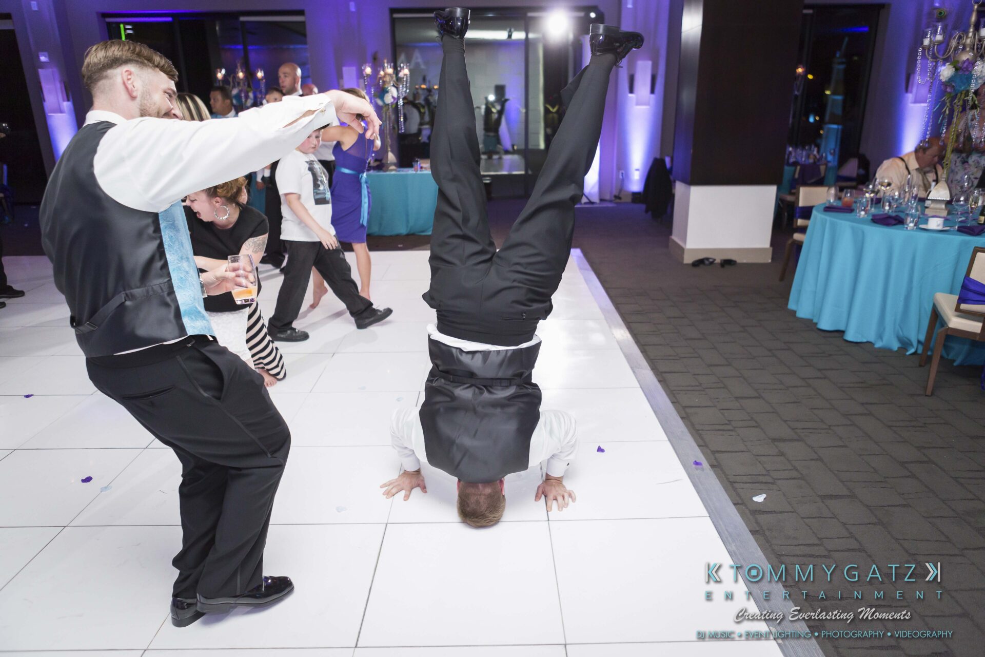 1211 - TOMMY GATZ ENTERTAINMENT A man in formal attire performs a handstand on a dance floor at a lively event, while others watch and dance in the background.